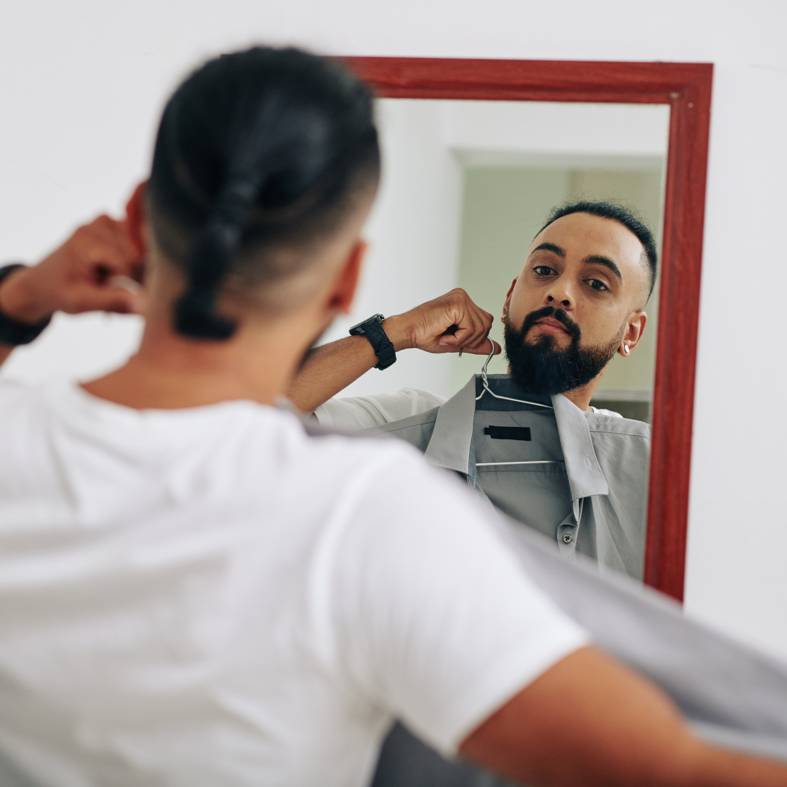 Homem com barba se olhando no espelho enquanto segura uma camisa em um cabide, avaliando sua escolha de roupa.