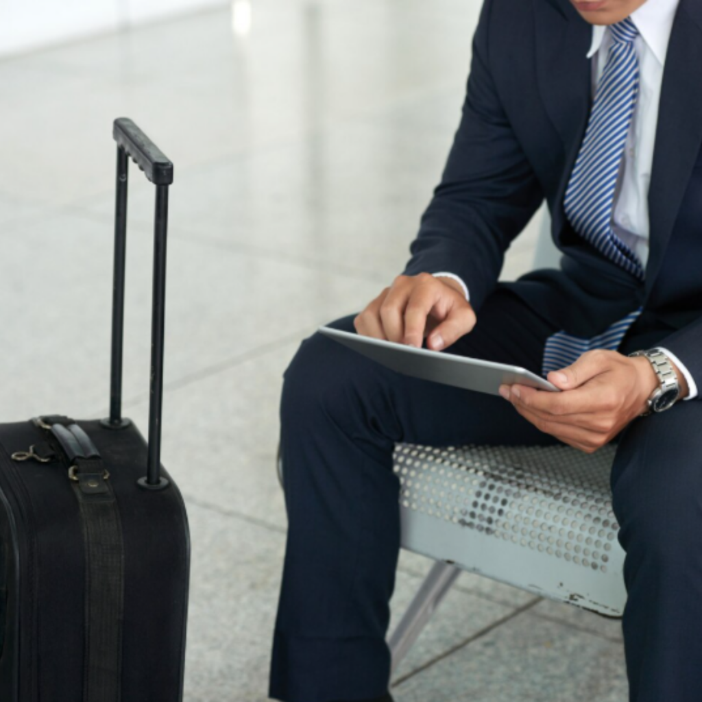 Homem de terno sentado em um aeroporto usando um tablet, com uma mala de viagem ao lado.
