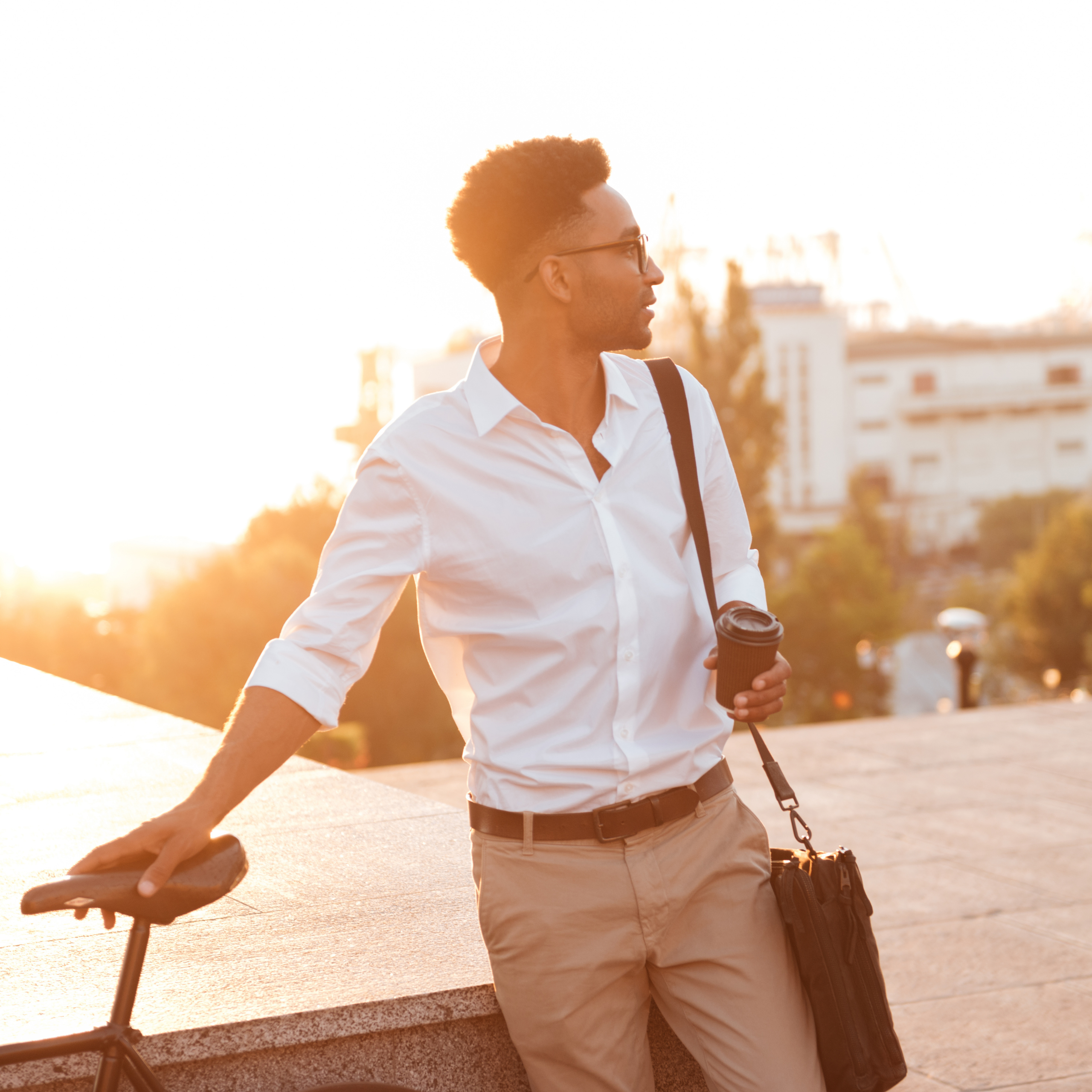 Homem vestindo camisa branca e calça clara, segurando um copo de café e apoiado em uma bicicleta, em ambiente urbano ao ar livre durante o pôr do sol.
